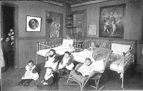 Historical photograph showing a group of children in a room with cribs and a painting on the wall.