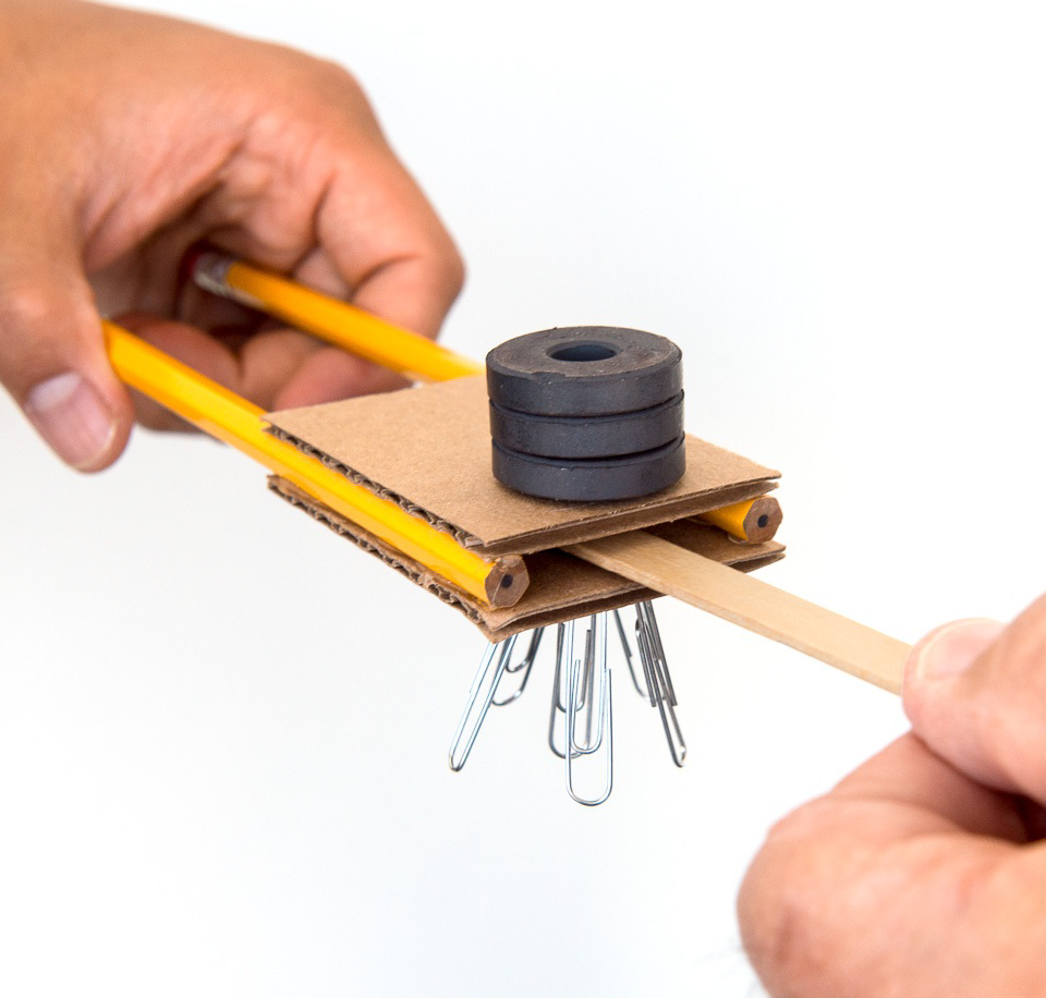 A setup showing magnets stacked on cardboard with pencils and paper clips to investigate magnetic shielding.