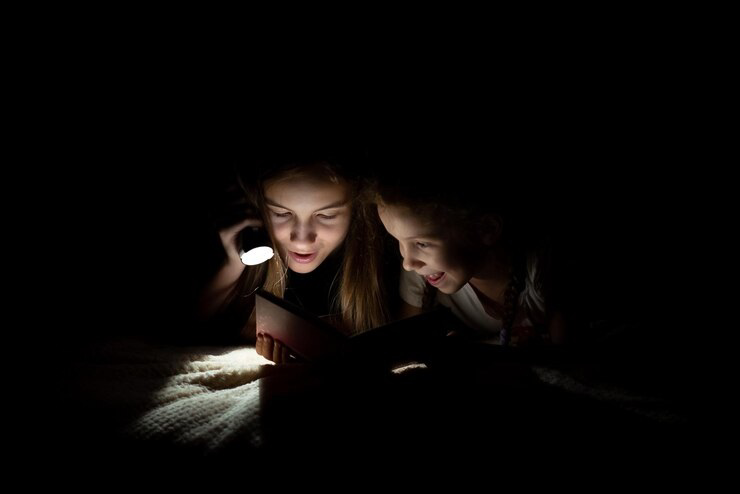 Two children reading a book in the dark using a flashlight.